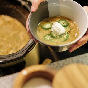 Hands holding a bowl of creamy crockpot white chicken chili topped with sour cream and jalapeños in front of a slow cooker