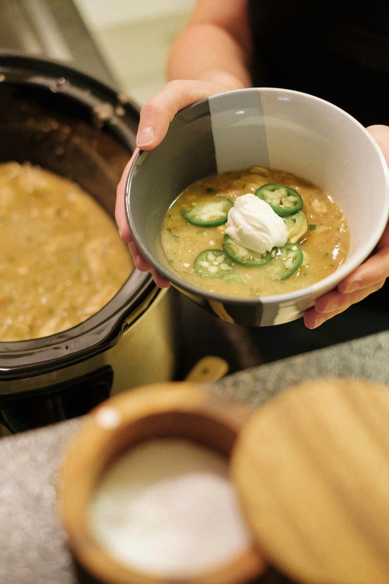 Hands holding a bowl of creamy crockpot white chicken chili topped with sour cream and jalapeños in front of a slow cooker