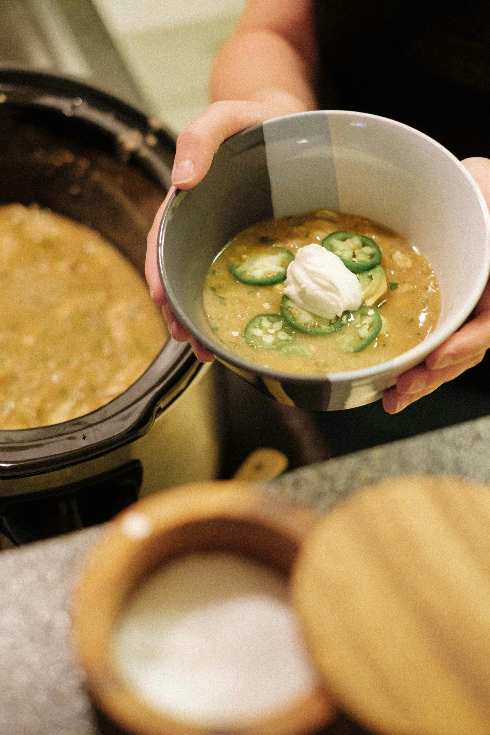 Hands holding a bowl of creamy crockpot white chicken chili topped with sour cream and jalapeños in front of a slow cooker